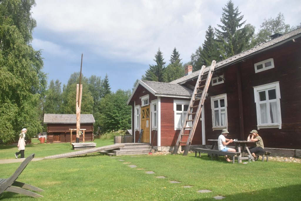 Kuvassa on punamultamaalissa oleva vanha maalaistalo, jonka pihalla on pöytä. Kaksi ihmistä kahvittelee pihapöydän ääressä. Kuvassa näkyy myöts vihreää nurmea ja pihaa sekä puita.