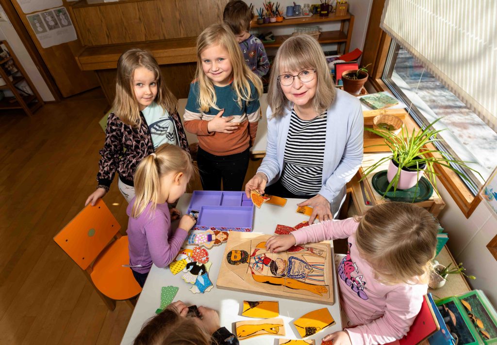 Kuvassa yksi aikuinen ja neljä lasta pelailee ja askartelee pöydän ääressä
