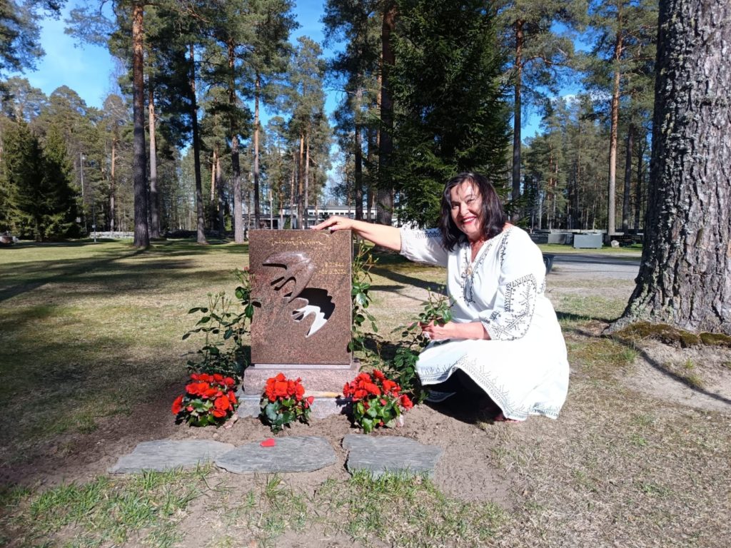 Nainen valkoisessa kirjotussa mekossa polvistuu hautakiven vieressä metsäisellä hautausmaalla ja hymyilee kameraan. Ruskeassa kivessä on perhosen muotoinen aukko, ja sen ympärille on asetettu punaisia kukkia ja ruusupensaita. Taustalla näkyy mäntyjä ja aurinkoinen kevätpäivä.