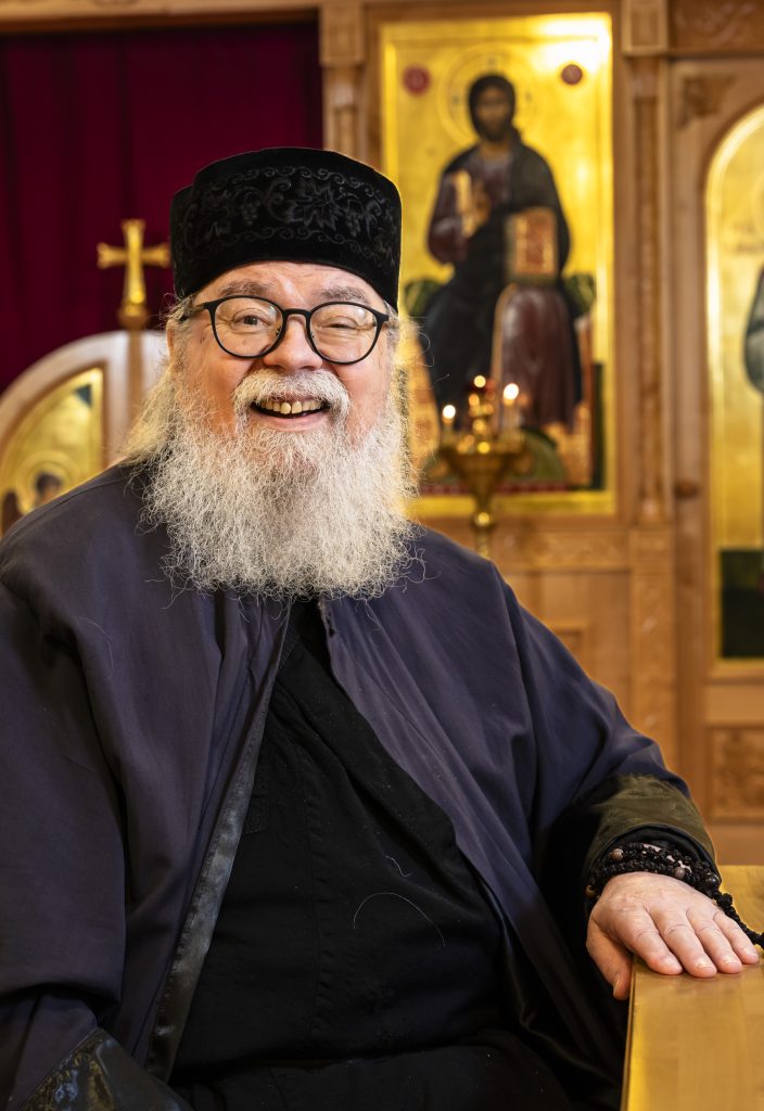 Hymyilevä ortodoksipappi istuu kirkossa ikonien edessä käsi puukaiteella. Hänellä on musta papinhattu, silmälasit ja pitkä harmaa parta, ja taustalla näkyy kynttilöitä sekä kultasävyisiä ikoneita.