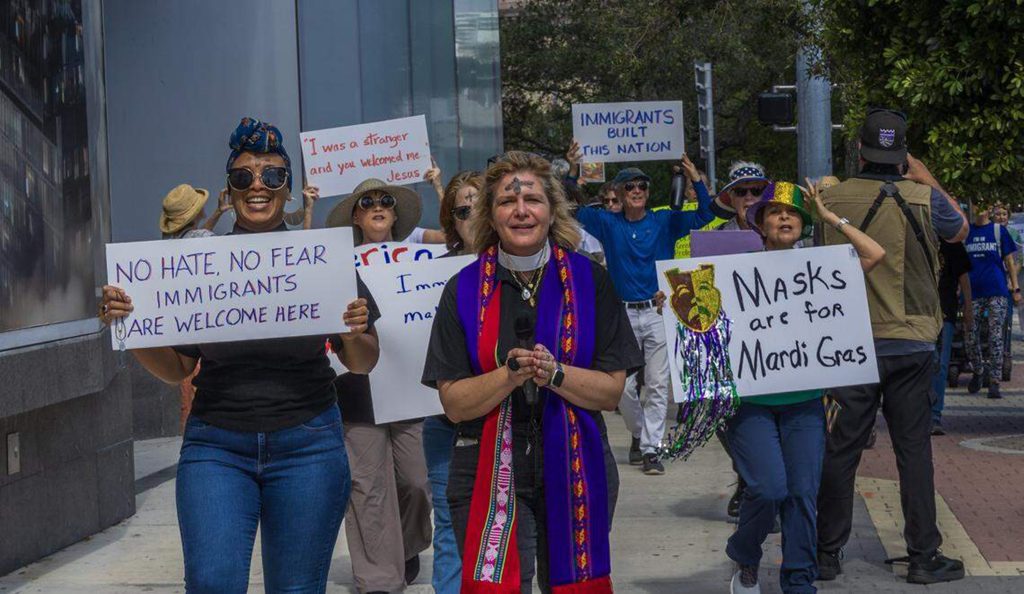 Joukko ihmisiä marssii kaupungin kadulla kantaen kylttejä, joissa ilmaistaan tukea maahanmuuttajille. Etualalla henkilö pitää kylttiä tekstillä ”No hate, no fear, immigrants are welcome here”, ja ryhmän keskellä kulkee papin asuun pukeutunut henkilö mikrofonin kanssa.