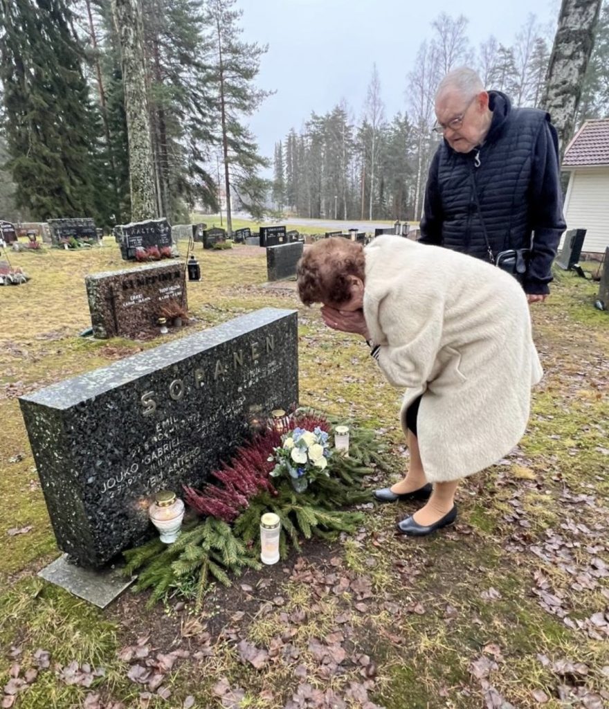Iäkkäät nainen ja mies hautausmaalla. Nainen on kumartuneena hautakiven puoleen ja asettanut juuri kukkakimpun kiven eteen. Häntä itkettää.
