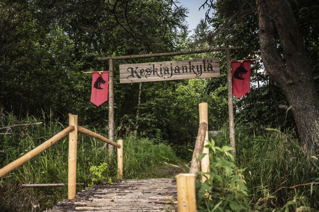 Kuvassa on puiset portaat metsän keskellä. Portaiden yläpäässä on puinen portti, jossa lukee Keskiajankylä