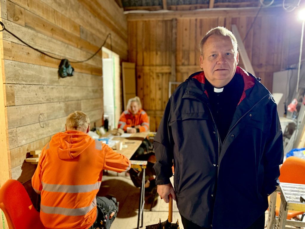 Pappi seisoo keskeneräisen puurakennuksen sisällä työvaatteisiin pukeutuneiden rakennustyöntekijöiden lähellä. Taustalla kaksi työntekijää istuu pöydän ääressä tauolla tai keskustelemassa, ja tilassa näkyvät puupinnat, rakennustarvikkeet ja työvalaistus.