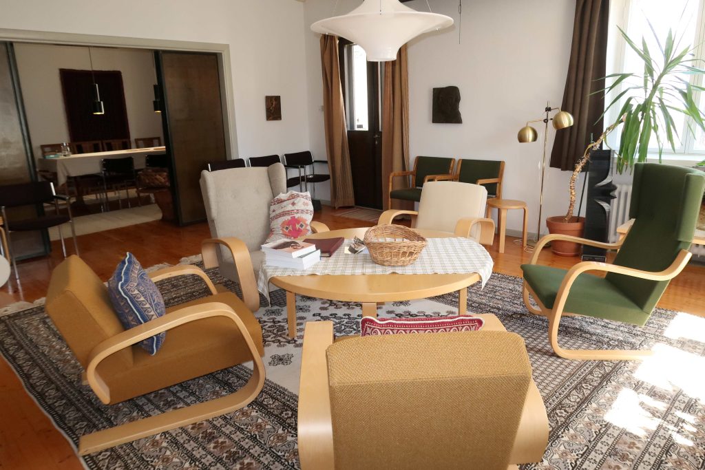 Kuvassa on huone, jonka keskellä on Artekin pöytä ja Artekin nojatuoleja. Katosta roikkuu Lokki-lamppu. Lattialla pöydän ja tuolien alla on iso matto. Kuvan oikeassa laidassa lattialla ruukussa on palmu.