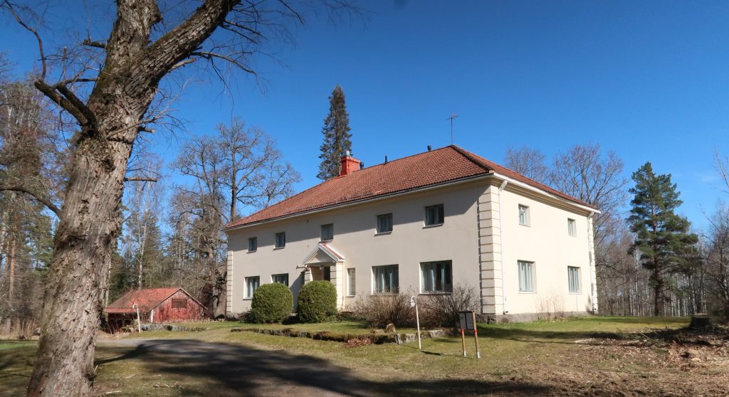 Kuvassa on vaalea, suuri kivirakennus jossa on punainen katto. Kuvassa näkyy myös puita ja pensaita sekä talon vasemmalla puolella puinen, punamultamaalinen, matala lato.