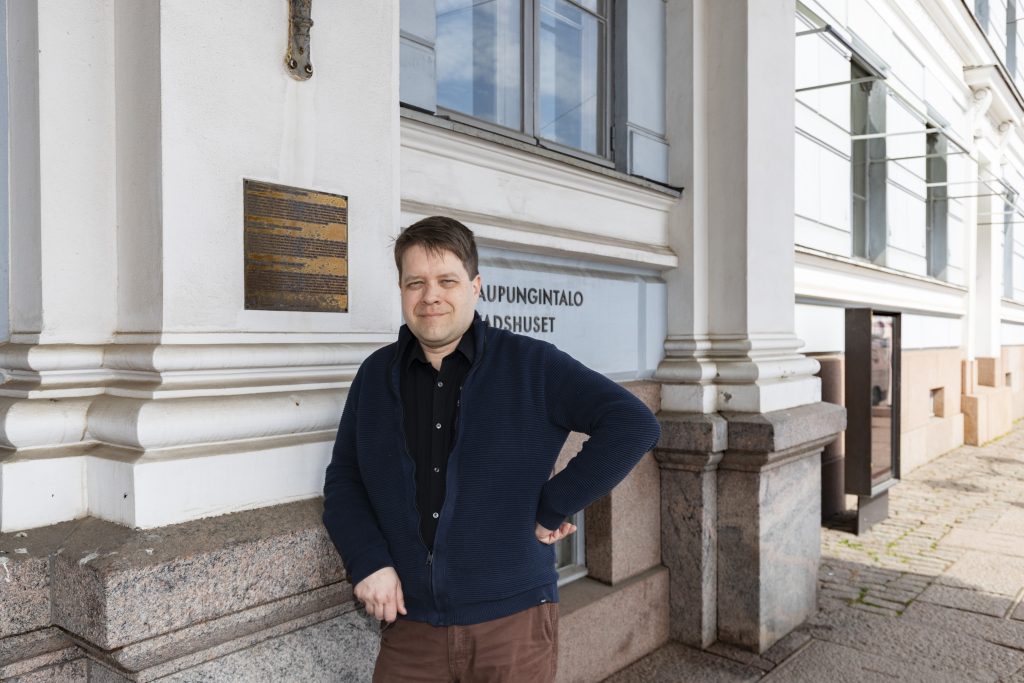 Henkilö seisoo vaalean kivirakennuksen julkisivun vieressä ja nojaa kädellään rakennuksen seinään. Taustalla näkyy suuria ikkunoita, pylväitä ja kaupungintalon kyltti rakennuksen seinässä.