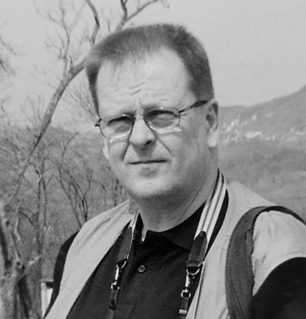 Mustavalkoinen muotokuva silmälasipäisestä henkilöstä ulkoilmassa. Hän katsoo kameraan ja hänen kaulassaan roikkuu kameran hihna. Taustalla näkyy lehdettömiä puita ja kauempana mäkinen maisema.