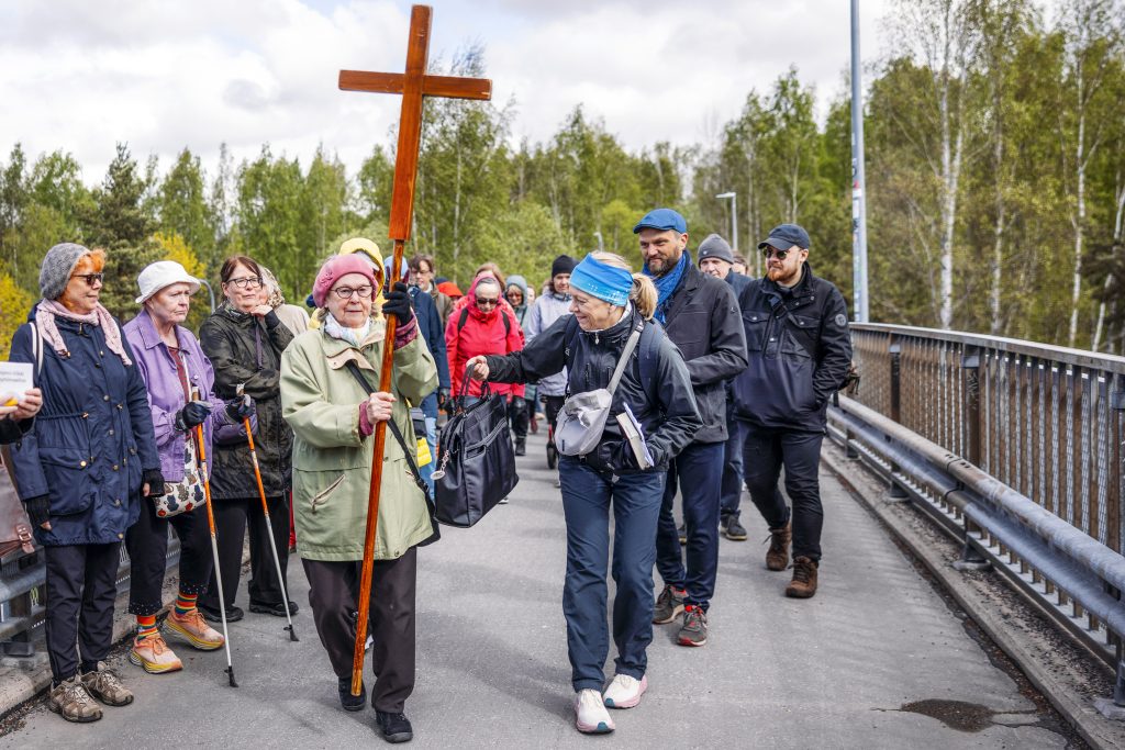 Ihmisryhmä kävelee sillalla metsäisen maiseman keskellä. Kulkueen eturivissä oleva henkilö kantaa suurta puista ristiä, ja muut osallistujat seuraavat perässä ulkoiluvaatteissa. Sillan kaiteet johdattavat katsetta kulkueen suuntaan.