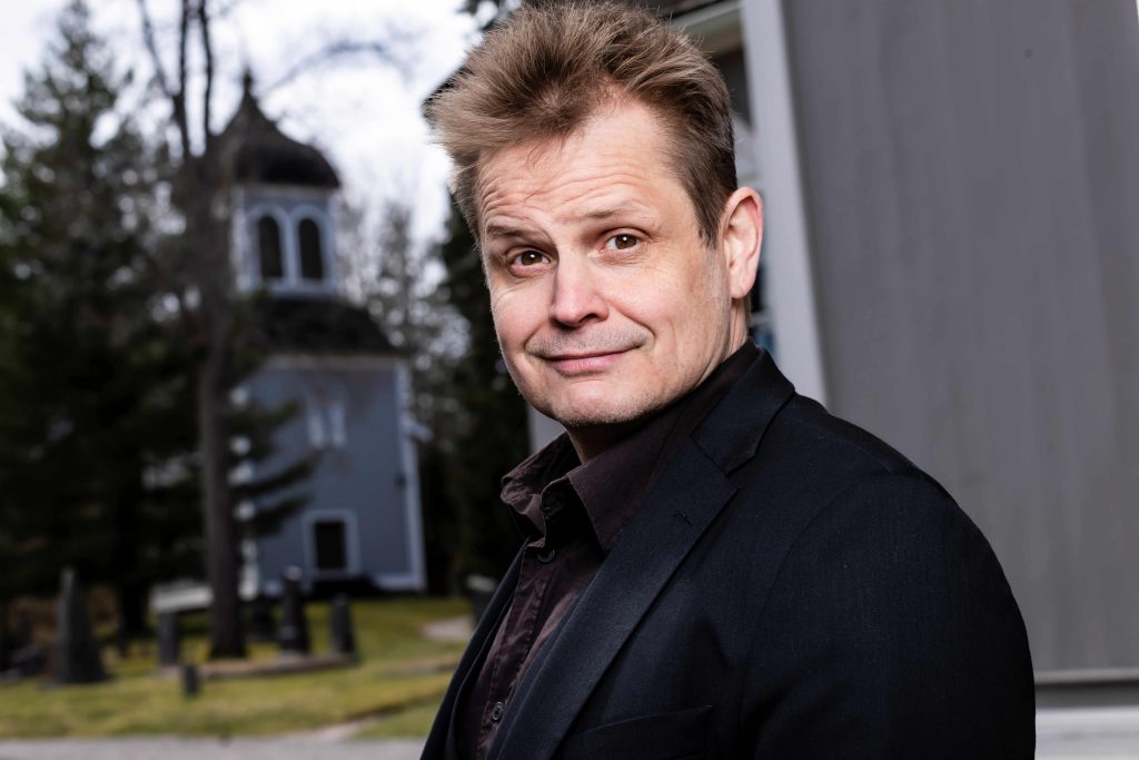 Hymyilevä mies seisoo kirkon pihalla puolilähikuvassa. Takana näkyy sumeasti puinen kellotapuli.