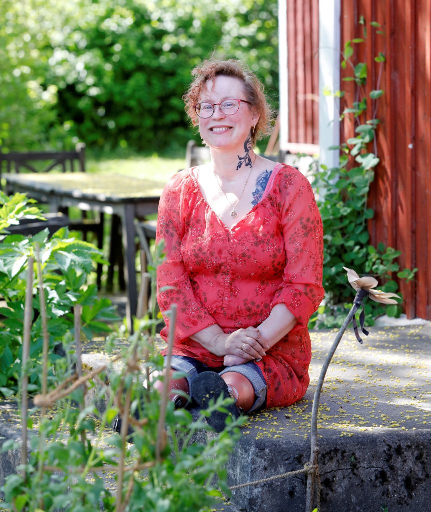 Kuvassa punatukkainen, punamekkoinen, silmälasipäinen nainen istuu omakotitalon portaalla kukkien ja kesäisen vehreyden keskellä.