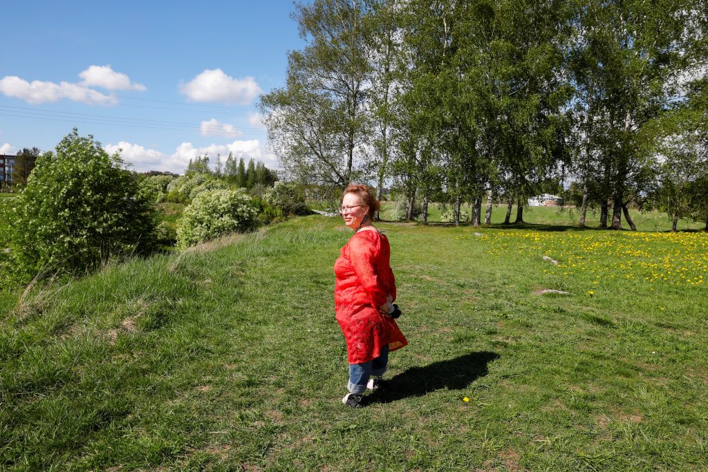 Kuvassa nainen punaisessa hameessa seisoo nurmikolla ja katsoo vasemmalle. Kuvassa näkyy myös vihreitä lehtipuita.