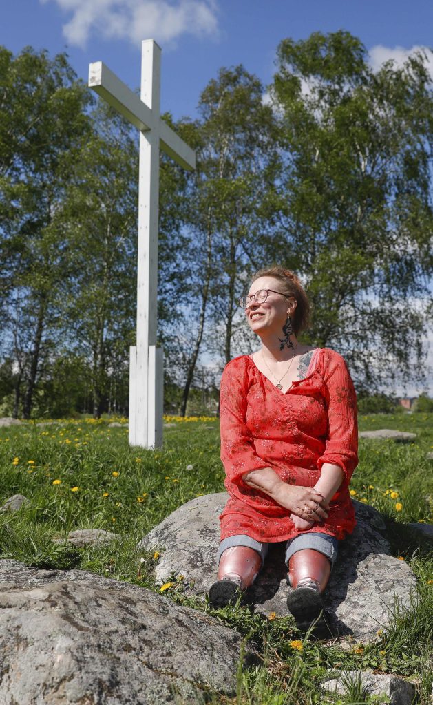 Kuvassa punamekkoinen silmälasipäinen nainen istuu kivellä nurmikolla ja katsoo silmät kiinni vasemmalle. Aurinko lämmittää kasvoja. Vieressä on korkea valkoinen, puinen risti.