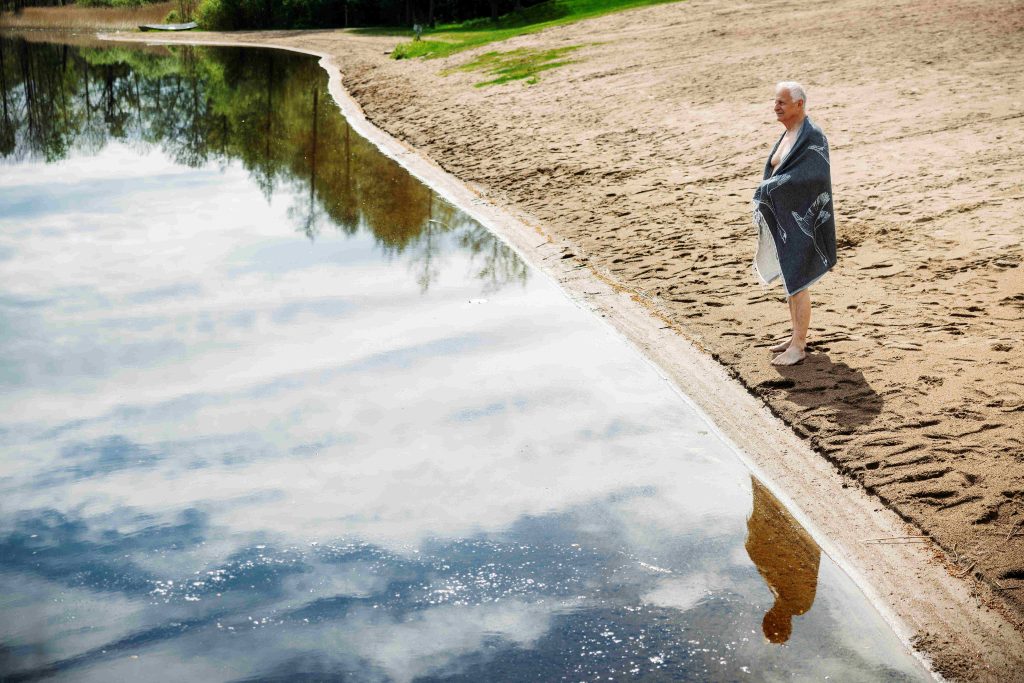 Kuvassa harmaahiuksinen mies seisoo pyyhe ympärillään järven rannassa hietikolla. Kuvassa näkyy järveä, josta heijastuu pilvien kuvajaisia ja miehen kuvajainen