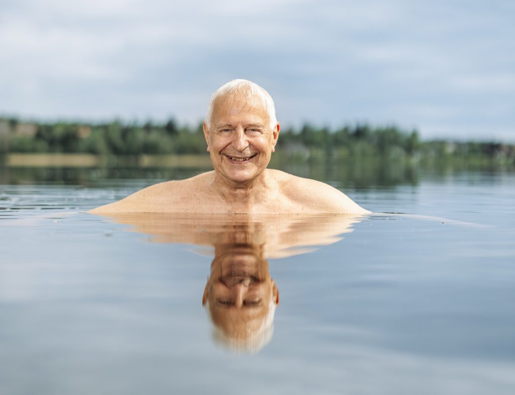 Kuvassa hymyilevän, harmaahiuksisen miehen pää pilkistää järvestä. Taustalla näkyy vastarannan metsikköä.