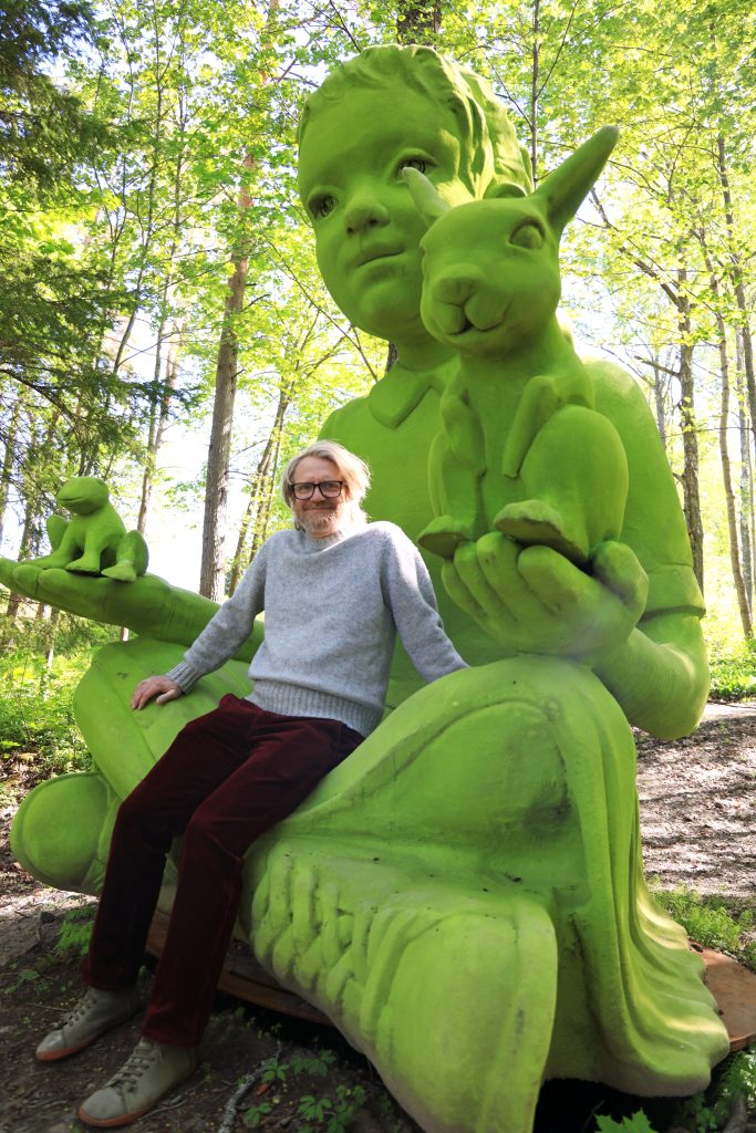 Kuvassa pitkähihaiseen, vaaleansiniseen paitaan ja mustiin housuihin pukeutunut mies istuu noin nelimetrisen, vihreän sammaljättipatsaan sylissä. Ympärillä on vihreää lehtimetsää.