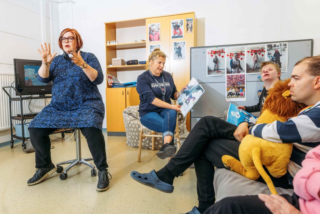 Hoitaja lukee satukirjaa kehitysvammaisten toimintakeskuksessa ja pappi viittoo sen viittomakielellä. Sohvalla istuu kuulijoita. Taustalla näkyy kirjahylly.