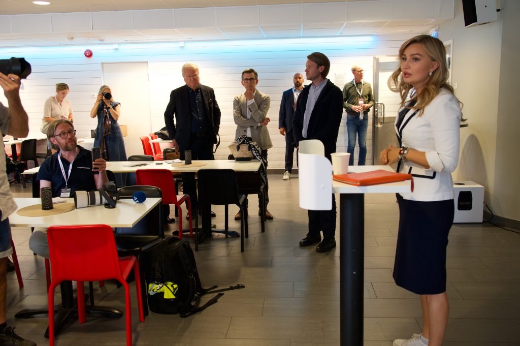Nainen puhuu korkean pöydän ääressä huoneessa, jossa joukko ihmisiä seuraa häntä. Paikalla on myös valokuvaajia ja kamerakalustoa.