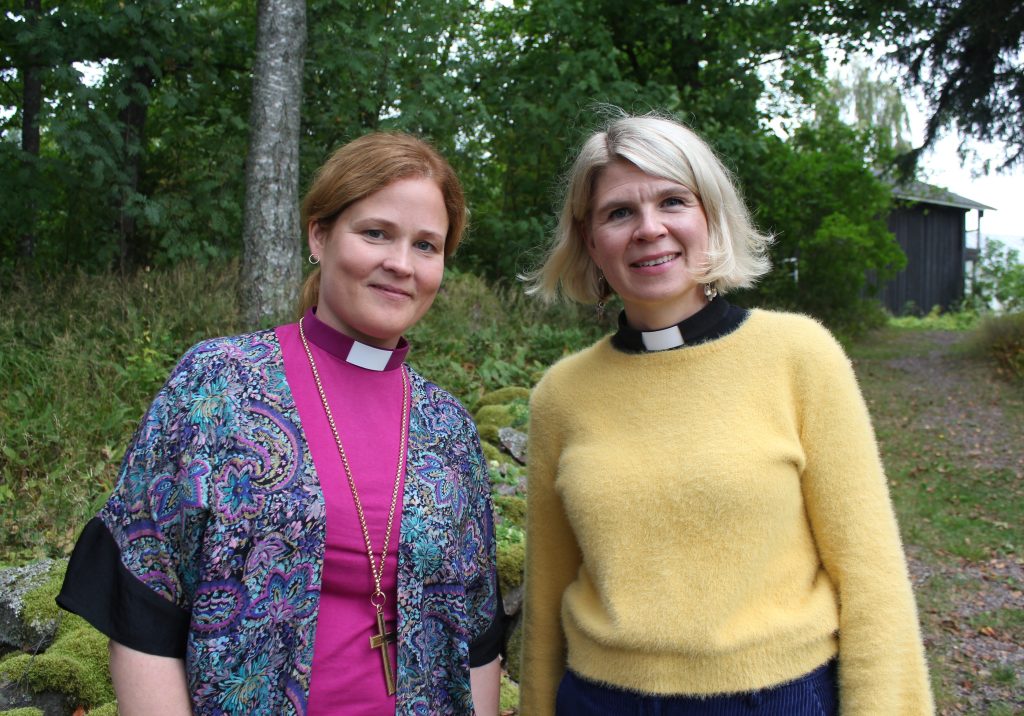 Tummatukkainen nainen violetissa piispanpaidassa, vierellään vaaleatukkainen keltaisessa paidassa. Luontoa taustalla.