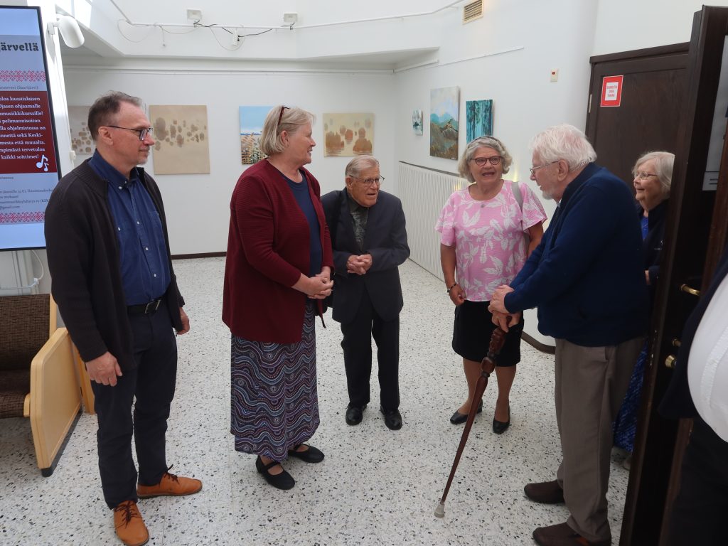 Ihmiset keskustelevat aula- tai galleriatilassa, taustalla seinällä näkyy maalauksia.