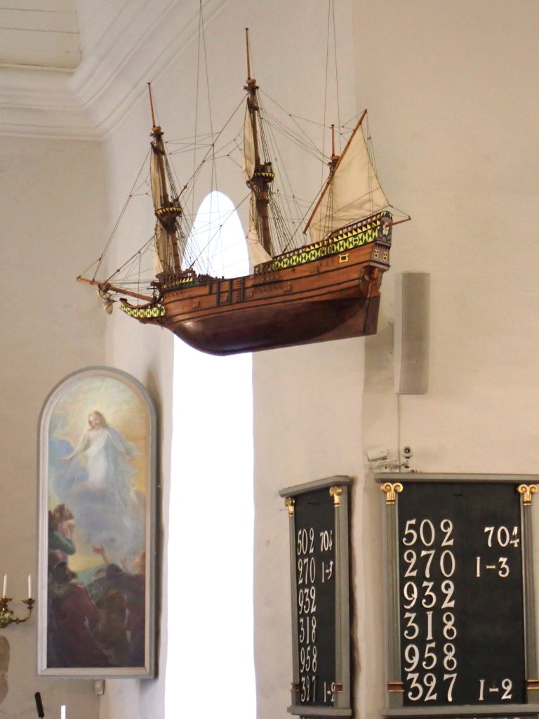Kirkossa otetussa kuvassa näkyy alttaritaulu, jossa on Jeesus sekä virsitaulu jossa on virsien numeroita ja katosta roikkuva purjelaivan pienoismalli