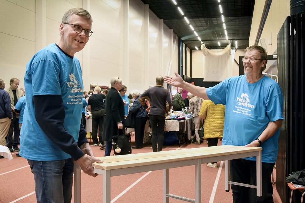 Kaksi sinisiin tapahtumapaitoihin pukeutunutta miestä esittelee vaaleaa puupenkkiä vilkkaassa sisätapahtumassa. Toinen pitää penkistä kiinni ja toinen viittoo sitä kohti. Taustalla ihmiset kiertävät myynti- ja esittelypöytien äärellä.