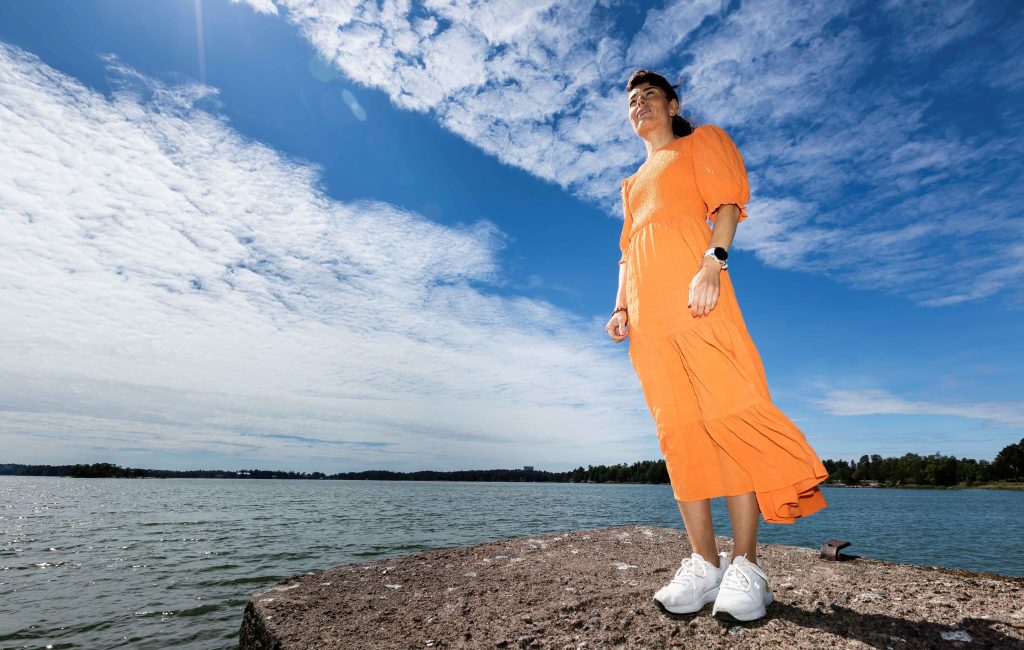 Kuvassa oranssiin pitkään mekkoon ja valkoisiin lenkkareihin pukeutunut nainen seisoo rantakalliolla. Taivaalla näkyy ohutta pilveä ja taustalla meri.