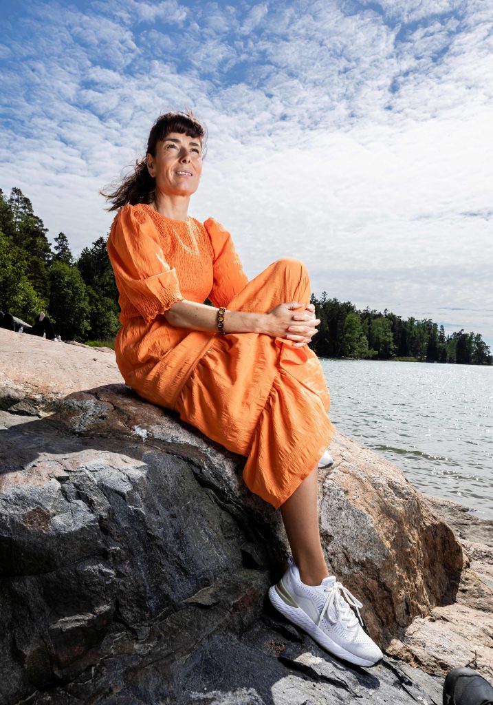 Kuvassa oranssiin pitkään mekkoon pukeutunut nainen istuu rantakalliolla. Taustalla näkyy merta ja metsää.