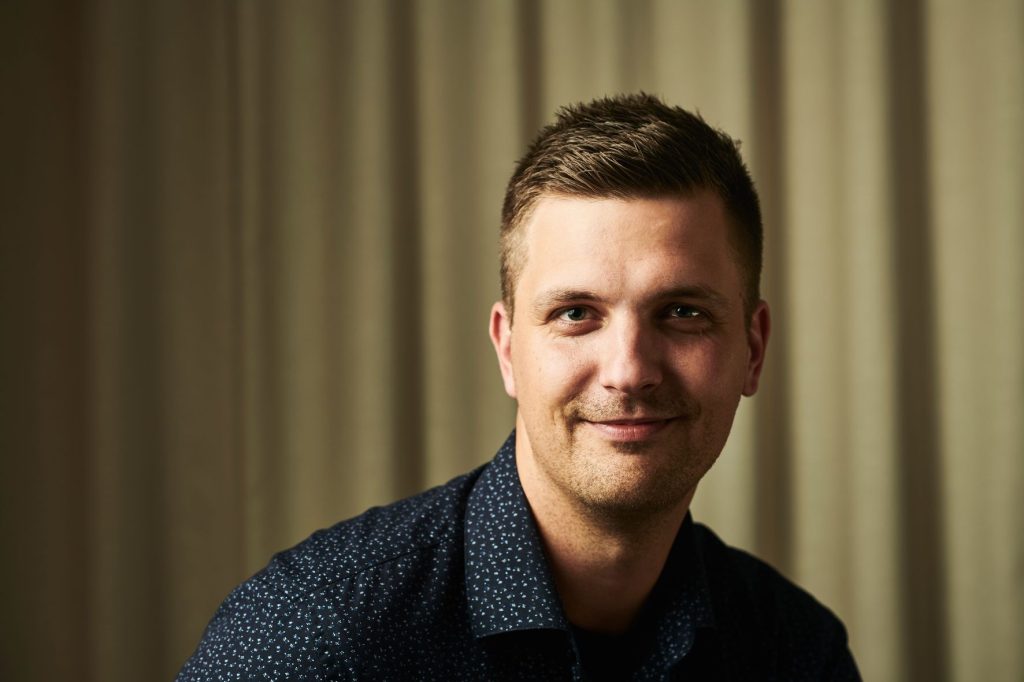 Lyhythiuksinen henkilö katsoo kameraan ja hymyilee kevyesti. Hänellä on tumma, pienikuvioinen kauluspaita, ja taustalla näkyy vaalea verho.