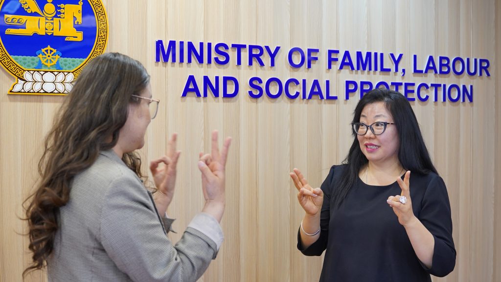 Puolilähikuvassa kaksi mongolialaista naista keskustelee viittomakielellä. Taustalla on vaalea puuseinä sekä sinisin kirjaimin englanninkielinen teksti: Ministry of family, labour and social protection
