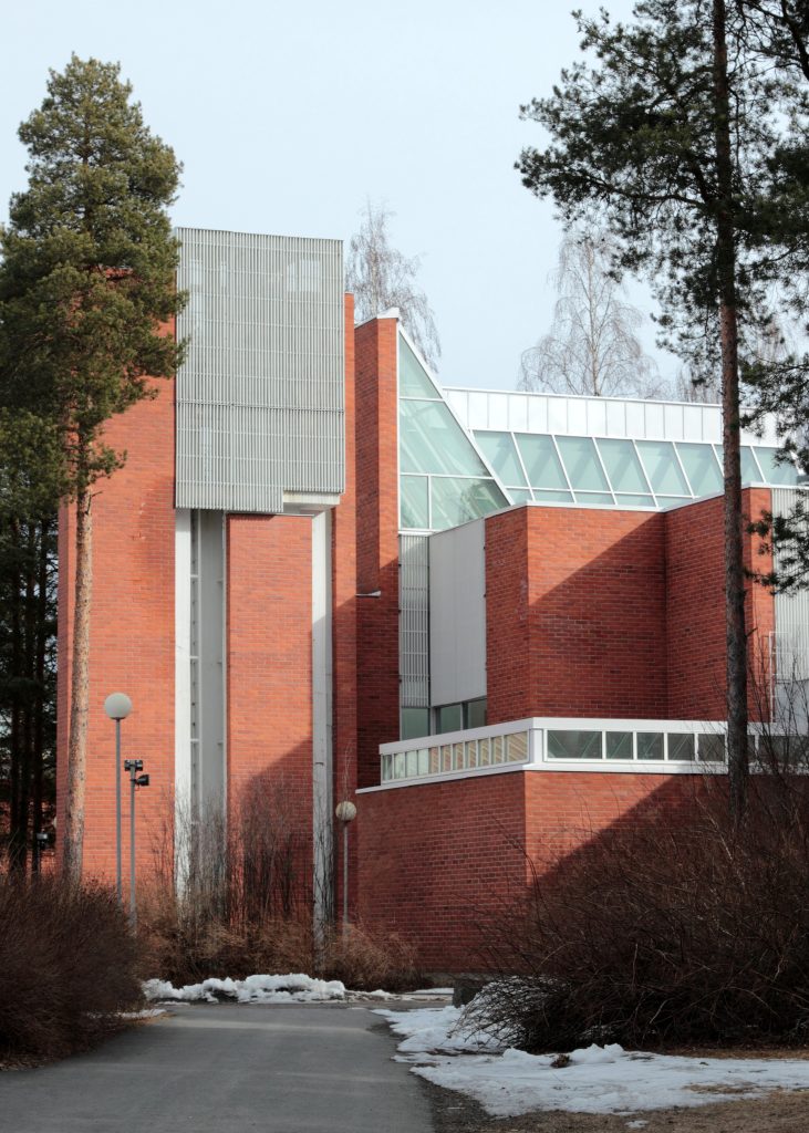 Punatiilinen, geometrisesti muotoiltu rakennus kohoaa mäntyjen keskellä. Julkisivussa vuorottelevat korkeat tiiliseinät, vaaleat metallisäleiköt ja suuret vinot lasipinnat. Etualalla kulkee osittain lumen reunustama pihatie.