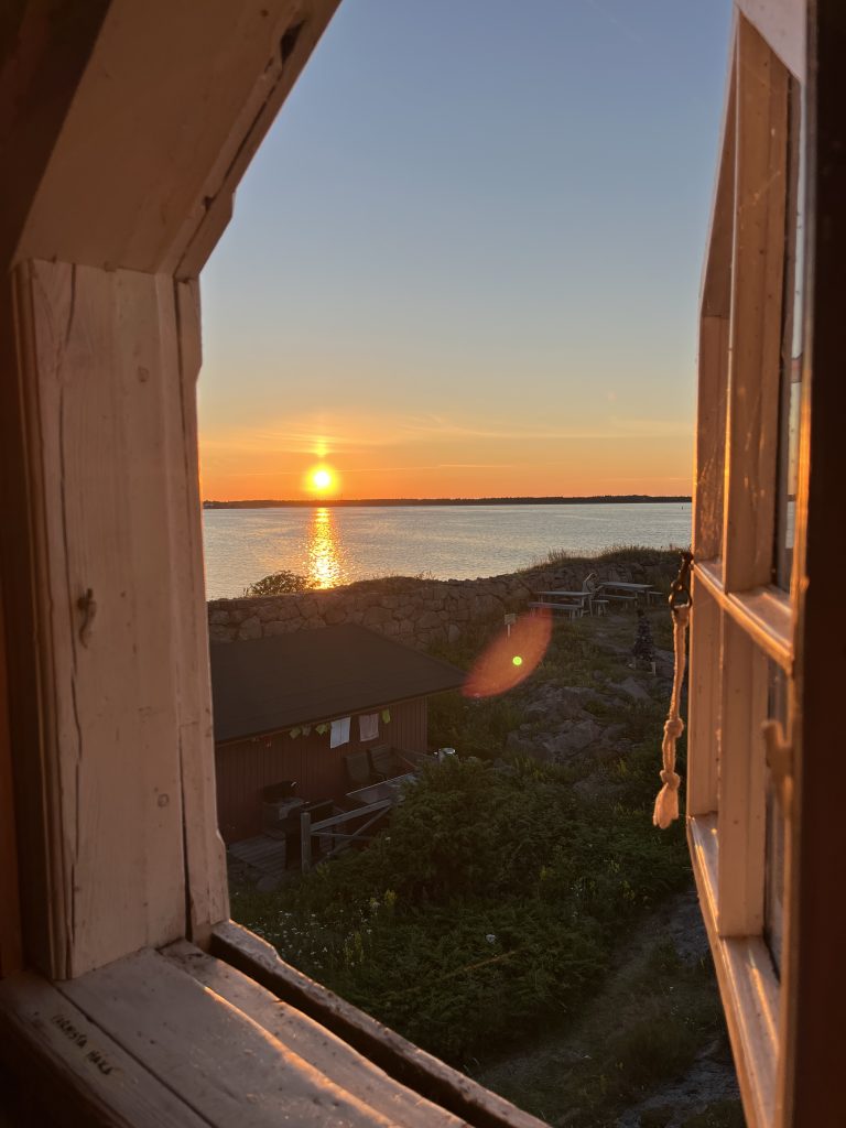 Kuvassa avonainen ikkuna josta näkyy auringonlasku merelle