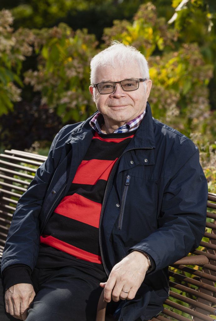 Valkohiuksinen, silmälasipäinen mies istuu puistonpenkillä ja katsoo hymyillen kameraan. Hänellä on tummansininen takki, punamustaraidallinen paita ja ruudullinen kauluspaita. Taustalla näkyy auringon valaisemia vihreitä pensaita ja kukintoja.