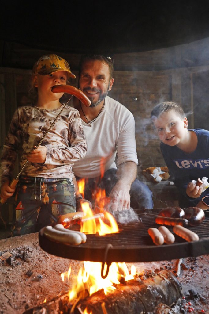 Aikuinen ja kaksi lasta paistavat makkaroita nuotiolla puisessa grillikatoksessa. Etualalla nuotion liekit kohoavat makkararitilän läpi. Keltalakkinen lapsi pitää makkaraa tikussa, aikuinen hymyilee keskellä ja toinen lapsi kurkistaa vierestä.