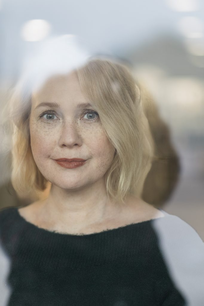 Lähikuvassa on vaaleahiuksinen nainen joka katsoo suoraan kameraan