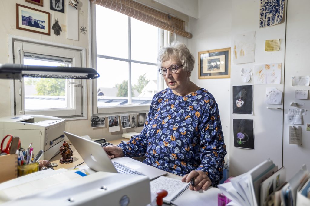 kuvassa sinikukalliseen paitaan pukeutunut vanhempi silmälasipäinen nainen kirjoittaa pöydän ääressä läppärillä, taustalla näkyy ikkuna
