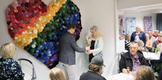 Järvenpään Jampan sydän sai seinälleen sateenkaaren värit − ”Usein istumme täällä tiuhassa kuin tupsut ryijyssä” Kirkkoherra Vesa Koivisto ojentaa kukat ryijyn ideoineelle Hanna Mäntyselle. Takana seinällä värikäs ryijy.