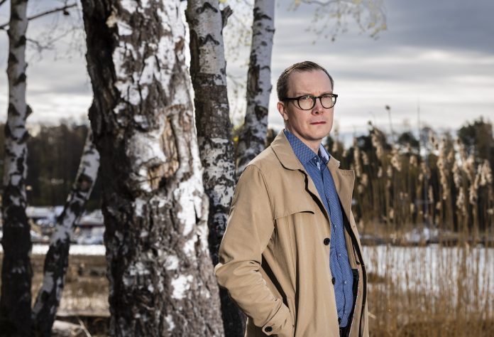 Mies beigessä takissa nojaa koivun runkoon. Takana näkyy vettä.