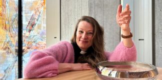 Vantaalla järjestetään oma kastepäivä 12–14-vuotiaille – ”Kasteessa liitytään suureen sanomaan” Nainen vaaleanpunaisessa neuleessa istuu pöydän ääressä, käsi nostettuna. Taustalla näkyy värikkäitä ikkunoita ja betoniseinää.