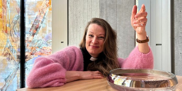 KM40230_marja eloranta 2_lpr Nainen vaaleanpunaisessa neuleessa istuu pöydän ääressä, käsi nostettuna. Taustalla näkyy värikkäitä ikkunoita ja betoniseinää.
