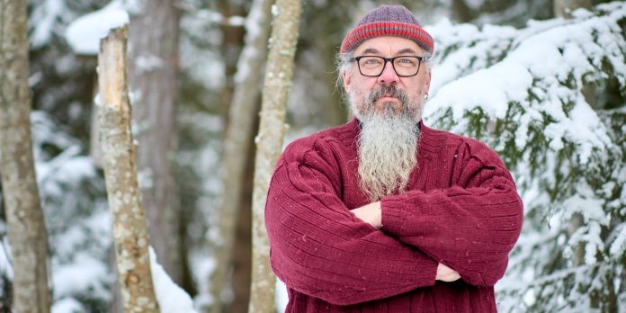 mies seisoo lumisessa metsässä, pitää käsiään puuskassa ja katsoo tuikeana kameraan