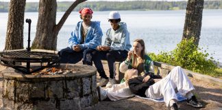 ”Pitäisi katsoa kokonaisvaikutuksia” − nuorisotyön säästöt herättävät huolta Nuoria istuu nuotiopaikalla järven rannalla, nauttien auringosta ja seurasta. Taustalla näkyy kaunis järvimaisema ja puita.