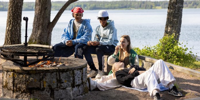 Nuoria istuu nuotiopaikalla järven rannalla, nauttien auringosta ja seurasta. Taustalla näkyy kaunis järvimaisema ja puita.