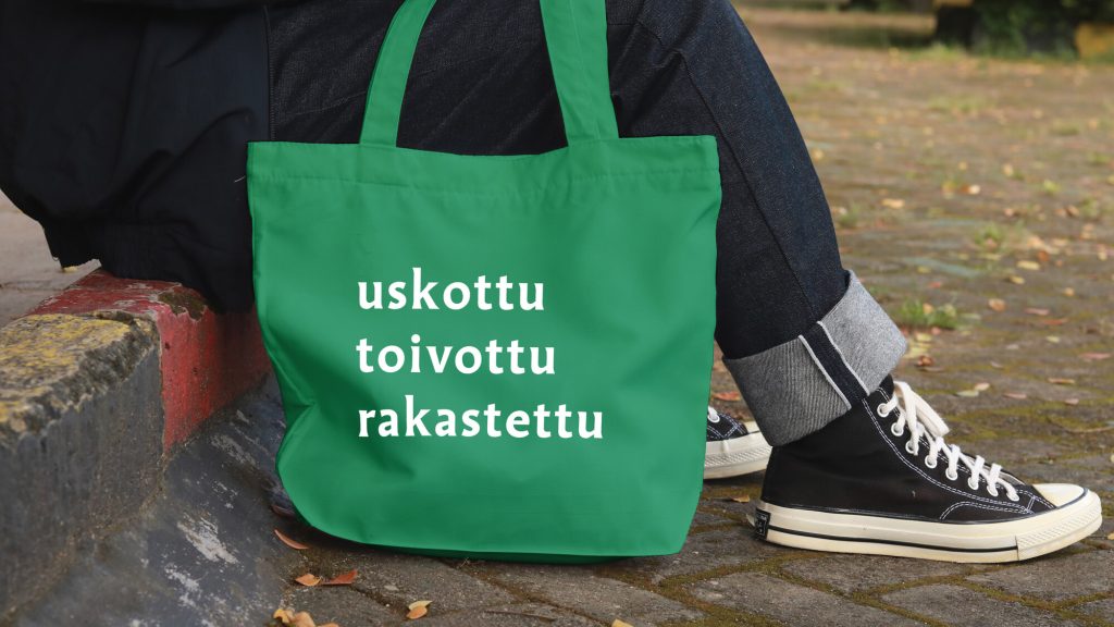 Vihreä kangaskassi, jossa on valkoiset sanat "uskottu", "toivottu" ja "rakastettu". Taustalla henkilö istuu kivetyksellä