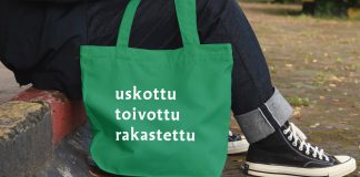 Vihreä kangaskassi, jossa on valkoiset sanat "uskottu", "toivottu" ja "rakastettu". Taustalla henkilö istuu kivetyksellä
