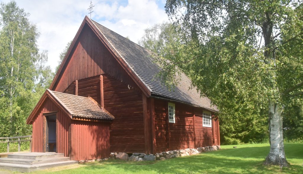Kuvassa on pieni, punamultamaalilla maalattu hirsikirkko vehreässä kesämaisemassa.