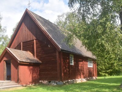 Kuvassa on pieni, punamultamaalilla maalattu hirsikirkko vehreässä kesämaisemassa.