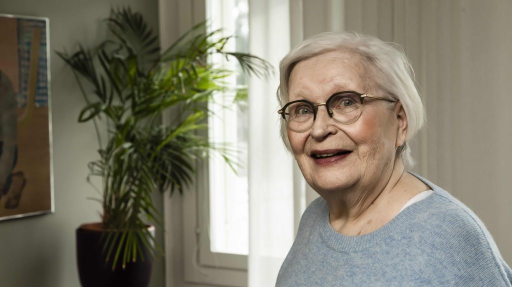 Kuvassa vanhempi, silmälasipäinen nainen katsoo puolilähikuvassa kameraan ja hymyilee. Taustalla näkyy palmu-huoneksvi.