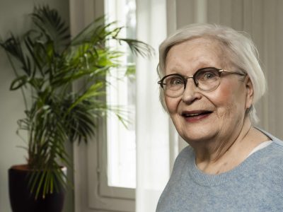 Kuvassa vanhempi, silmälasipäinen nainen katsoo puolilähikuvassa kameraan ja hymyilee. Taustalla näkyy palmu-huoneksvi.