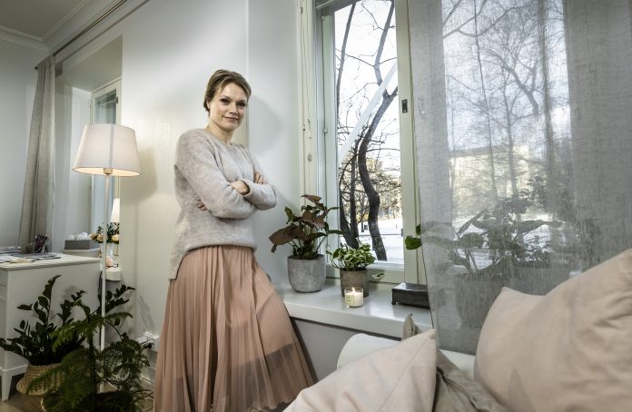 Nainen nojaa olohuoneessa ikkunan pieleen. Ikkunalaudall palaa kynttilä.